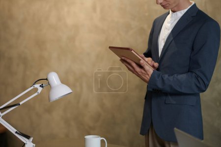 Photo for Cropped image of young businessman standing at office desk and reading report on digital tablet - Royalty Free Image