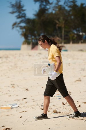Téléchargez les photos : Jeune homme avec seau en plastique marchant sur la plage et ramassant les déchets - en image libre de droit