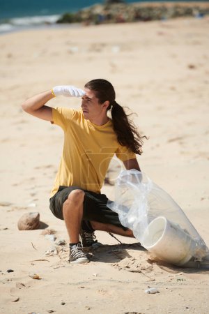 Téléchargez les photos : Jeune homme portant des gants lors de la collecte des déchets sur la plage dans un sac en plastique - en image libre de droit