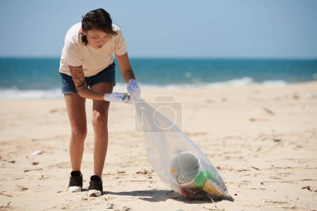 Téléchargez les photos : Jeune femme nettoyer la plage de la mer, ramasser les déchets dans un sac en plastique - en image libre de droit