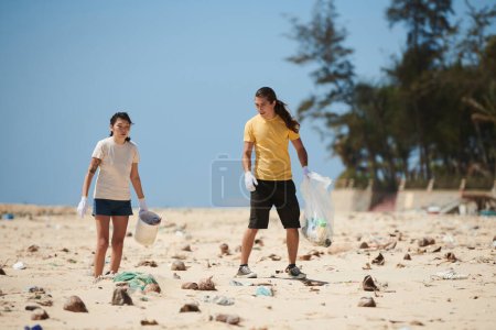 Téléchargez les photos : Jeune couple ramassant débris et déchets laissés par les touristes sur la plage de la mer - en image libre de droit