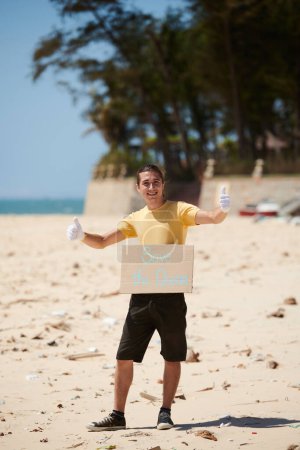 Téléchargez les photos : Portrait de militant heureux excité debout sur une plage sale avec sauver la plaque de l'océan et montrant pouces levés - en image libre de droit
