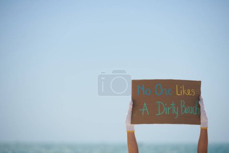 Téléchargez les photos : Mains de bénévoles tenant personne n'aime la plaque de plage sale contre le ciel bleu - en image libre de droit