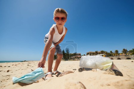 Foto de Niña en gafas de sol limpiando la playa del mar de mascarillas usadas - Imagen libre de derechos