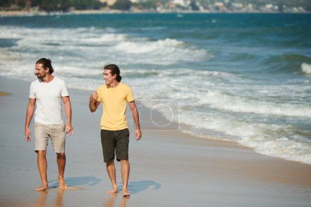 Téléchargez les photos : Amis masculins positifs appréciant marcher sur la plage de la mer par une journée ensoleillée - en image libre de droit
