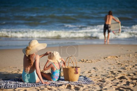 Photo for Mother and daughter sitting on blanket and looking at father surfing in sea - Royalty Free Image