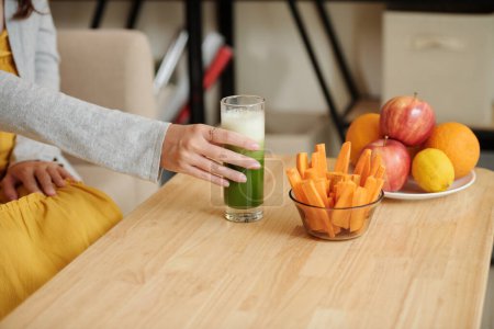Téléchargez les photos : Femme enceinte ayant smoothie vert, bâtonnets de carotte et fruits frais pour collation - en image libre de droit