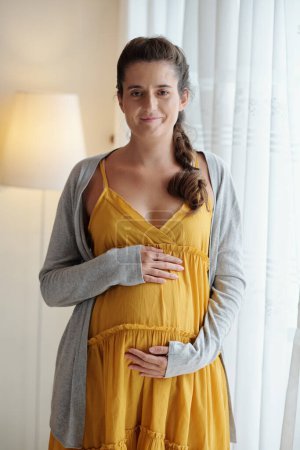 Foto de Retrato de una mujer embarazada sonriente tocando el vientre y mirando a la cámara - Imagen libre de derechos