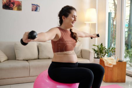 Foto de Mujer embarazada alegre sentada en la pelota de fitness y haciendo ejercicio con pequeñas mancuernas - Imagen libre de derechos