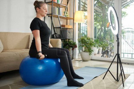 Foto de Entrenador personal en línea sentado en la pelota de fitness y explicando cómo entrenar con pesas - Imagen libre de derechos