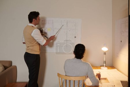 Photo for Engineer pointing at blueprint on wall when discussing details with colleague - Royalty Free Image