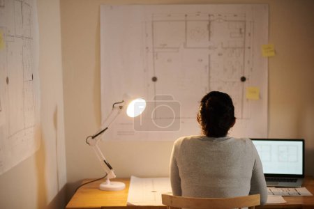 Téléchargez les photos : Architecte féminine pensive regardant le plan de construction accroché au mur au-dessus de son bureau dans une pièce sombre, vue de dos - en image libre de droit