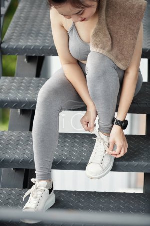 Foto de Ajuste mujer joven en ropa deportiva sentado en los escalones y atar cordones de zapatos antes de correr por la mañana - Imagen libre de derechos