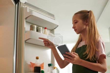 Photo for Preteen girl with shopping list on smartphone checking shelves in fridge - Royalty Free Image
