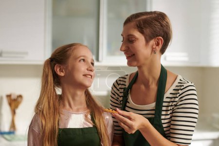 Téléchargez les photos : Mère et fille souriantes dégustant des biscuits qu'elles ont cuits ensemble à la maison - en image libre de droit