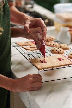 Téléchargez les photos : Mains de mère et fille décorer maison en pain d'épice avec glaçage rouge - en image libre de droit