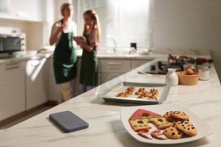 Téléchargez les photos : Assiette avec biscuits au pain d'épice sur le comptoir de la cuisine, mère et fille parlant en arrière-plan - en image libre de droit