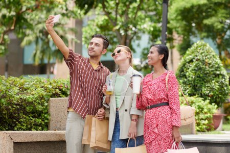 Téléchargez les photos : Jeune homme prenant selfie avec des amis après avoir fait du shopping ensemble le vendredi noir - en image libre de droit