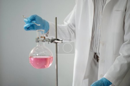 Foto de Científico de laboratorio añadiendo un poco de polvo en frasco con líquido rosa - Imagen libre de derechos