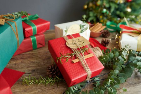 Téléchargez les photos : Petit cadeau de Noël enveloppé dans du papier rouge sur la table à côté de cadeaux pour les membres de la famille - en image libre de droit