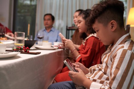 Téléchargez les photos : Les enfants passent du temps à faire défiler les médias sociaux sur les smartphones lors du dîner familial - en image libre de droit