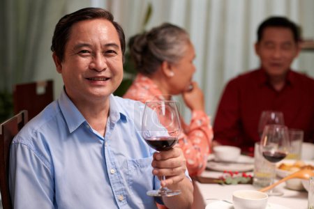 Photo for Portrait of cheerful Asian man toasting with glass of wine when sitting at dinner table with family members - Royalty Free Image