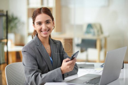 Photo for Portrait of cheerful young businesswoman with smartphone in hand sitting at office desk - Royalty Free Image