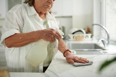 Téléchargez les photos : Image recadrée d'une femme âgée désinfectant le comptoir de cuisine avec du détergent - en image libre de droit