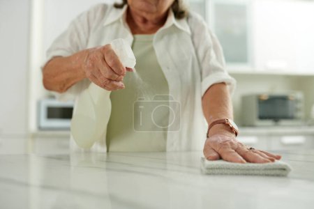 Téléchargez les photos : Senior femme pulvérisation de détergent sur le comptoir de cuisine lors du nettoyage de la maison - en image libre de droit