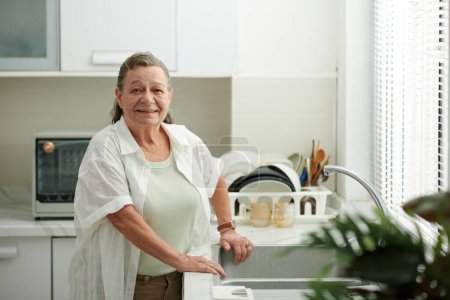 Foto de Retrato de mujer mayor sonriente de pie en el fregadero de la cocina - Imagen libre de derechos