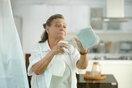 Foto de Ventana de limpieza mujer anciana con detergente en aerosol y paño suave - Imagen libre de derechos