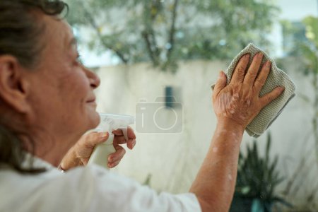 Foto de Imagen recortada de la mujer anciana limpiando la puerta de vidrio con detergente en aerosol - Imagen libre de derechos