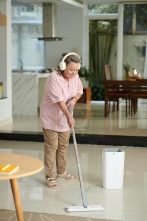 Téléchargez les photos : Femme âgée écoutant de la musique dans les écouteurs lors de l'essuyage du sol dans sa maison - en image libre de droit