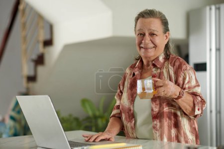 Téléchargez les photos : Portrait de femme âgée heureuse buvant du thé et vérifiant les e-mails sur ordinateur portable - en image libre de droit