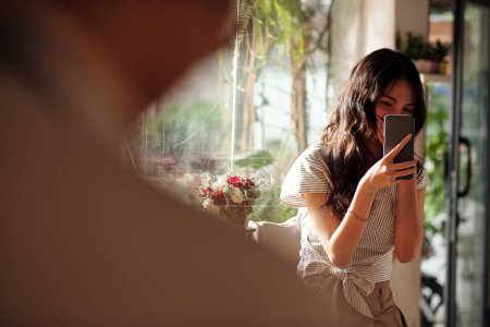 Foto de Sonriente joven enamorada fotografiando secretamente su enamoramiento en coffeeshop - Imagen libre de derechos