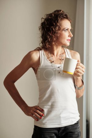 Foto de Contemplando mujer en blanco tanque superior beber taza de café y mirando a través de ventana del apartamento - Imagen libre de derechos