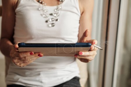 Téléchargez les photos : Mains de femme en débardeur blanc fumant la cigarette et lisant l'e-book ou l'article sur la tablette - en image libre de droit