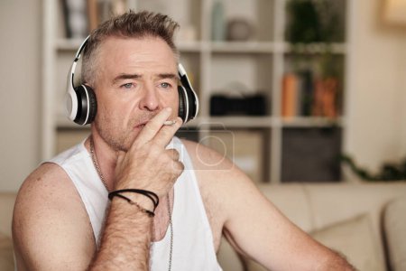 Photo for Portrait of middle-aged man smoking cigarette and listening to music or podcast in headphones - Royalty Free Image