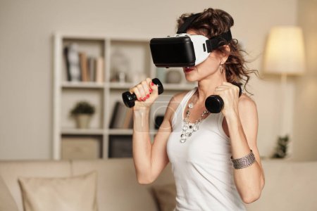 Photo for Portrait of middle-aged woman in virtual reality headset exercising with dumbbells - Royalty Free Image