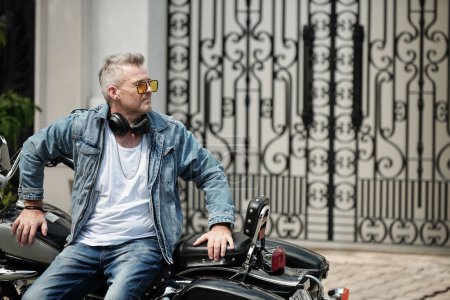 Téléchargez les photos : Cool homme mature élégant en jeans et veste en denim apprendre à moto et détourner les yeux - en image libre de droit
