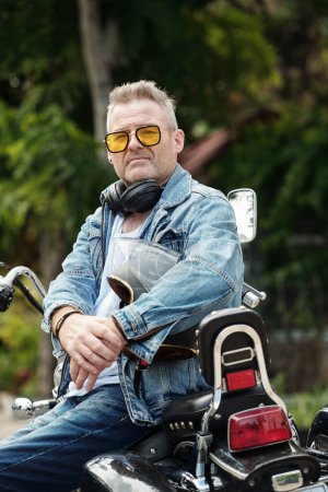 Foto de Retrato de hombre maduro sentado en motocicleta con casco en las manos y mirando a la cámara - Imagen libre de derechos
