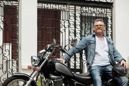 Téléchargez les photos : Pensive homme mûr appuyé sur la moto en attendant petite amie à l'extérieur - en image libre de droit