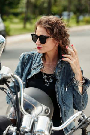 Téléchargez les photos : Femme mûre glamour dans des lunettes de soleil assis sur la moto et regardant loin - en image libre de droit