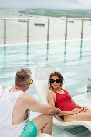 Foto de Mujer madura en traje de baño rojo y gafas de sol relajándose en el chaise-lounge y mirando a su marido que habla - Imagen libre de derechos
