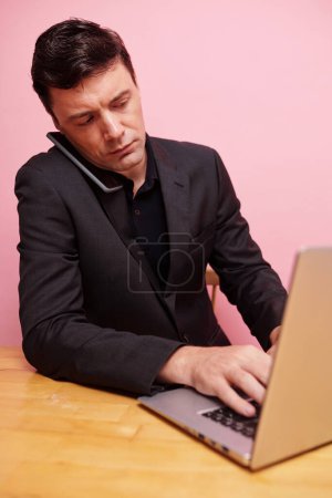 Téléchargez les photos : Homme d'affaires sérieux parlant au téléphone et tapant sur un ordinateur portable - en image libre de droit