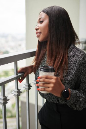 Photo for Young elegant businesswoman standing on office balcony with cup of take out coffee and enjoying city view - Royalty Free Image