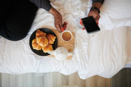 Foto de Mujer tomando té con croissants en la cama mientras revisa las redes sociales en el teléfono inteligente, ver desde arriba - Imagen libre de derechos