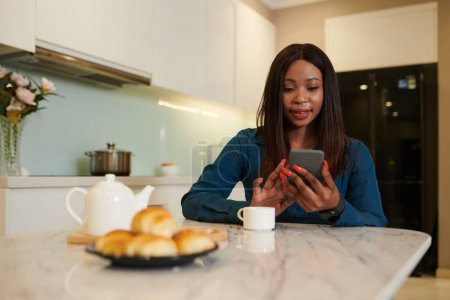 Foto de Mujer de negocios negro comprobar las notificaciones en el teléfono inteligente al tomar café con croissant en la mañana - Imagen libre de derechos