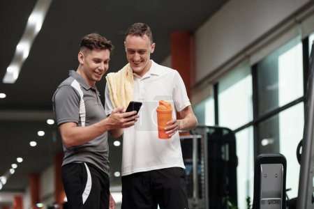 Téléchargez les photos : Sportsman montrant drôle de vidéo sur smartphone à un ami quand ils s'entraînent dans la salle de gym - en image libre de droit