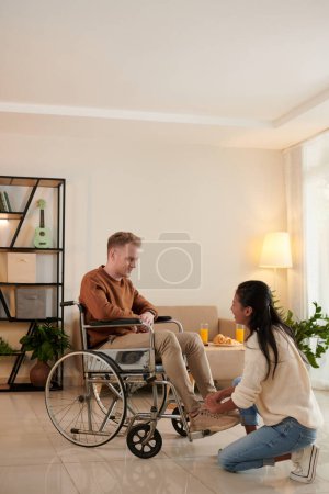 Téléchargez les photos : Petite amie attentionnée attachant lacet de beyfriend assis dans un fauteuil roulant - en image libre de droit